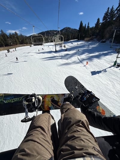Two snowboarders ride a chairlift uphill at a ski resort. The sunny, snow-covered slopes feature visible tracks and scattered skiers below. A chairlift line runs parallel, supported by metal towers. A mix of trees flanks the mountainside. The snowboarders are seen from a close perspective, showcasing their boots strapped onto their boards. One snowboard is black and the other is multicolored with a design that says 'Custom'.