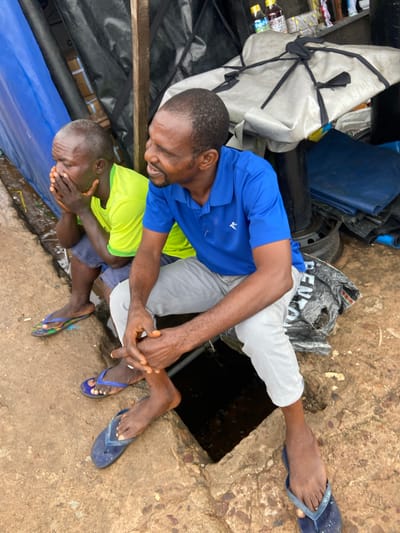 Two men, likely of Nigerian descent, are interacting outdoors in what appears to be an informal or somewhat cluttered setting. One man, wearing a blue shirt and light-colored pants, is kneeling or crouching, reaching towards the other man. The second man, younger and wearing a bright green shirt and shorts, is lying down and has his hands covering his face. His body is oriented in a way that suggests he might be distressed or in pain.
The ground is a rough, concrete-like surface, with some dirt and debris visible. There are various objects in the background, including what looks like tarpaulins, bundles wrapped in white material, a blue plastic container, and a discarded tire. A black plastic bag with white lettering is also present. The lighting suggests it might be daytime, but the overall scene appears somewhat dim or overcast. The presence of a visible opening in the ground, possibly a well or drain, is a significant detail in the setting. The man in the blue shirt is wearing dark flip-flops, and the younger man is wearing patterned flip-flops. The scene appears to be in Ogbunike, Nigeria. The overall emotion conveyed is one of concern or distress, particularly from the younger man's posture.