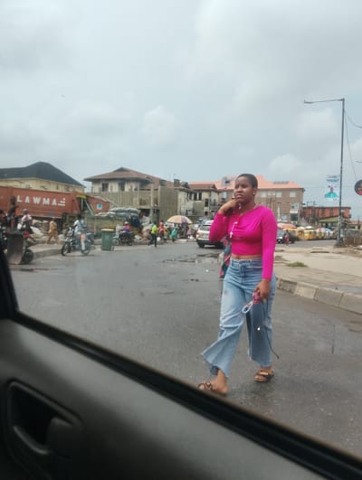 A young woman in a pink crop top and loose-fitting blue jeans walks along a street in a busy urban area, possibly a market. She is holding a phone to her ear and looking to her left. She wears sandals and a rosary around her neck. In the background, several buildings are visible, including some under construction. A large orange truck with "LAWMA" written on its side, likely a waste management vehicle, is parked on the side of the road, surrounded by refuse. Motorbikes and a few cars are also present, suggesting it is daytime. The sky is overcast, indicating cloudy weather. The overall scene depicts a bustling street in Alagbede, Nigeria, with everyday life unfolding amidst commercial activity and waste management operations.