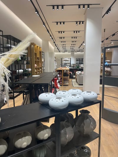 The image captures the interior of a retail store, likely a home decor or furniture store, in Santiago de Surco, Peru. The store features a modern design aesthetic with an emphasis on clean lines and neutral colors.
In the foreground, a black metal display shelf is stocked with decorative objects. These include white, sphere-shaped vases with perforated patterns, as well as darker, irregularly-shaped pottery. The shelf’s contents suggest a curated selection of design-forward items.
The background reveals a long, narrow retail space with rows of shelves and displays filled with various merchandise. The store's interior is illuminated by numerous track lights mounted on the ceiling. The lighting design contributes to a bright and inviting atmosphere.
The perspective of the image implies that it was taken from within the store, looking down the central aisle. There are no people visible in the immediate foreground, but there are suggestions of possible customers in the far background. The store appears to be well-organized and presented, appealing to customers seeking stylish home furnishings and decor.