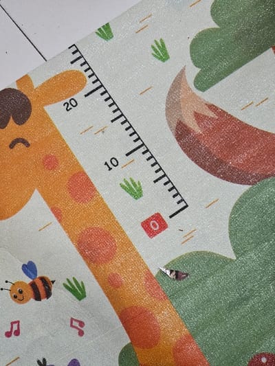 This is a close-up, oblique view of a patterned, glittery-textured mat, reportedly located in Muka, Indonesia. The mat features various cartoon illustrations against a light, off-white background. Prominent elements include a segment of an orange giraffe's body with darker orange spots and part of its brown head, positioned on the left. Adjacent to the giraffe is a vertical measuring scale marked in black, displaying numerical values "10" and "20," indicating increments. To the right, a reddish-brown fox tail with a lighter tip is visible. Below the giraffe's head, a yellow and black striped bee with blue wings is depicted. Other illustrations scattered across the mat include small green plant shapes, pink musical notes, small orange dashes, and a red square containing a white '0'. A small, irregularly shaped, metallic-silver object, approximately 0.5 cm in length, rests on the mat near a green hill-like illustration. No individuals or active interactions are present in the frame.