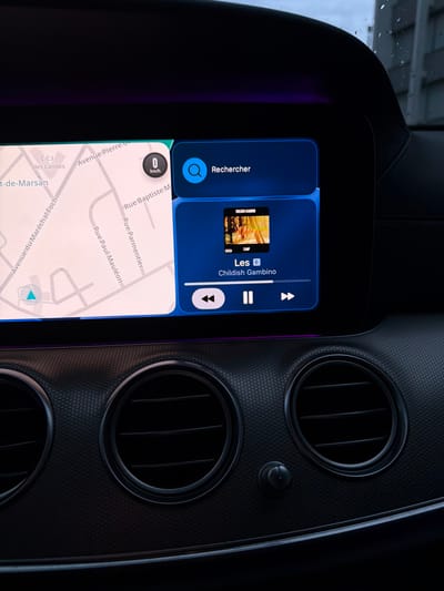 The image displays the interior of a vehicle, focusing on the central infotainment system and part of the dashboard. The infotainment screen is divided into two main sections. On the left, a digital map shows street layouts within the city of Mont-de-Marsan, France, with visible street names including "Avenue du Maréchal Foch," "Rue Paul Mauléon," "Rue Parmentier," and "Avenue Pierre." A digital speedometer overlay shows "0 km/h." On the right, a media player interface is active, displaying album art for "CAMP" by "Childish Gambino," with the song title partially visible as "Les." Control icons for previous, play/pause, and next are present, along with a "Rechercher" (Search) bar. Below the screen, the dashboard features a textured surface, two circular air vents with horizontal slats, and a small, cylindrical control knob. A faint purple ambient light strip is visible along the base of the screen, just above the dashboard. The upper portion of the image shows a blurred view through the windshield, indicating an overcast sky with possible rain droplets on the glass, suggesting an exterior environment during daytime. No individuals are visible.