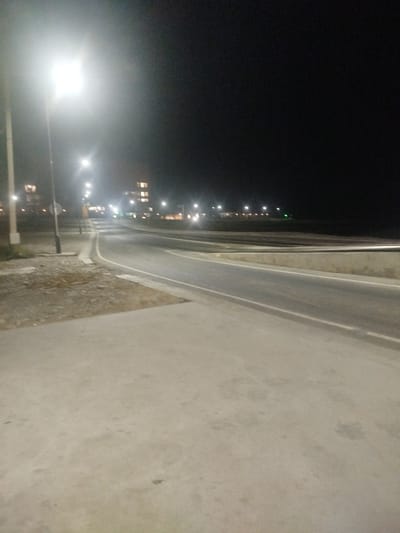 The image depicts a road at night, illuminated by streetlights. The road appears to be an asphalt surface with white lane markings, and it curves to the left in the foreground. To the left of the road, there is a concrete walkway and an area with gravel and dirt. In the background, several streetlights line the road, and further in the distance, buildings with illuminated windows are visible. The sky is dark, indicating it is nighttime. The scene appears to be an outdoor setting, possibly on the outskirts of a town or city, given the presence of both the road infrastructure and the distant buildings. There are no people visible in the image. The lighting suggests it might be a clear night, as the streetlights are bright and distinct. The location context suggests this is the bypass road 84 in Kyrgyzstan.
