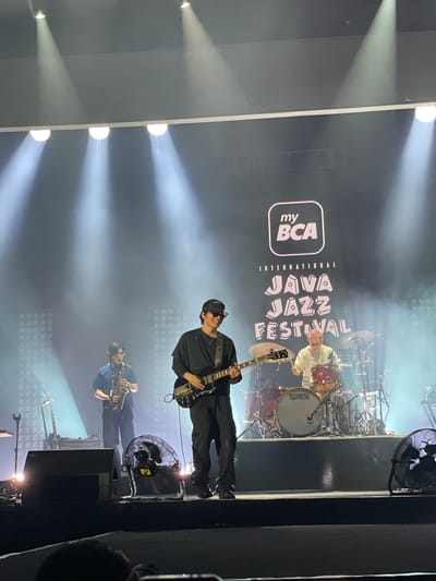 The image captures a live performance at the my BCA International Java Jazz Festival in Salembaran, Indonesia. The stage is lit with spotlights, creating a bright atmosphere. A guitarist, dressed in all black, stands center stage. Below him, a saxophone player in a blue shirt is seen. Behind the guitarist, a drummer is visible behind his drum kit. There is a logo for the festival, "my BCA International Java Jazz Festival," on a screen behind the musicians. A large electric fan sits on the stage as well. The scene conveys the ambiance of a live music event.