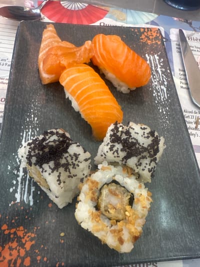 The image captures a meal consisting of sushi and maki rolls, presented on a dark grey rectangular plate with white and orange decorative speckles. This scene is situated on a paper placemat at a restaurant table, likely in Clamart, France, given the French text visible on the placemat.
On the plate, there are six pieces of sushi. Three pieces are salmon nigiri, each featuring a vibrant orange slice of raw salmon draped over a compact bed of white rice. The remaining three pieces are maki rolls. Two of these rolls are covered generously with small, dark black spherical seeds. The third maki roll, located at the bottom right, reveals a textured, possibly fried or tempura-battered, interior filling and is topped with numerous golden, crispy flakes.
The immediate environment visible includes a silver spoon partially at the upper left corner and a silver knife positioned at the upper right. A dark blue or black object, possibly a glass or bottle, is partially visible behind the knife. The paper placemat beneath the plate features various printed details, including text in French, such as "Bière" and "Alcool," and decorative illustrations, including a prominent red fan design and subtle light blue parasols. No people, specific actions, or direct interactions are visible within the frame.