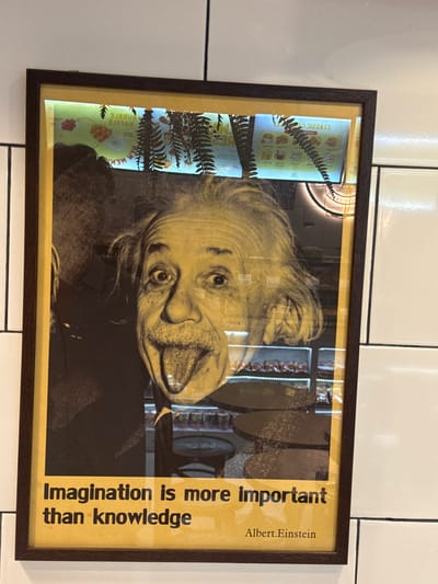 This media file is a framed print featuring a black and white photograph of Albert Einstein sticking his tongue out. The print is oriented vertically, with the top of Einstein's head pointing towards the left. To the left of Einstein's face, there is a vertical strip of what appears to be a product display or advertisement, featuring images of packaged goods and foliage, possibly ferns. The text "CRYZ21C CO" is visible on one of the packages, though it is reversed and difficult to read clearly.
To the right of Einstein's image, there is text printed vertically on a yellow background. The text reads: "Imagination is more important than knowledge". Below this, it says "Albert. Einstein". The overall scene suggests the print is displayed indoors, possibly on a tiled wall. The lighting appears to be artificial, creating reflections on the glass covering the print. The setting is described as Mai Khao, Thailand, but there are no specific visual cues within the print itself to confirm this location. The emotion conveyed by Einstein's expression is playful and unconventional.