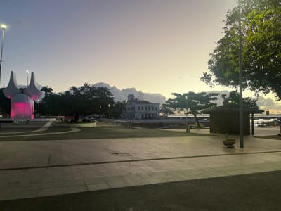 The image captures Praça Cairu in Salvador, Brazil, at either dawn or dusk. The scene is dominated by a paved public plaza extending into the midground. On the left, a large, white modernist monument with two upward-curving elements features a central cylindrical section illuminated with pink light. Adjacent to this monument, a mature tree shades a circular bench where two individuals are seated. A tall streetlight stands near the monument.
On the right side of the plaza, a small, dark-wood kiosk with a sloped roof is positioned beneath another large tree, near an active streetlight. A unique sculpture composed of stacked, rounded stone elements rests on the pavement in front of the kiosk.
In the background, a multi-story historic white building with a red-tiled roof and a prominent central tower, identified as the Mercado Modelo, overlooks the Baía de Todos-os-Santos. A pier with benches extends into the water from the waterfront. The sky displays a gradient of purple, orange, and yellow tones with scattered clouds, consistent with early morning or late evening lighting conditions. Artificial illumination from streetlights and the monument is visible.