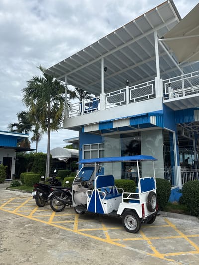 In Don Sak, Thailand, a white and blue electric vehicle is parked in a yellow-lined parking spot on a concrete surface. The vehicle has two rows of blue seats and a spare tire mounted on the rear. To its left, two motorcycles are parked, one black and the other black with red accents. Behind the vehicles, a multi-story building features a blue and white facade with large glass windows. Palm trees are visible on the left side of the image. The sky is overcast.
