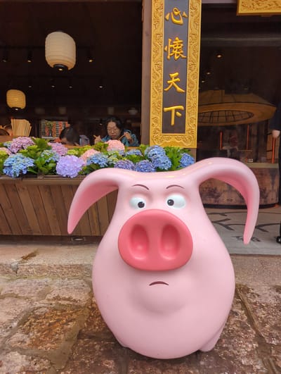 A pink pig sculpture with wide eyes, a large snout, and downturned ears is positioned in the foreground on a cobblestone path. Behind the sculpture, a wooden planter filled with vibrant pink, blue, and purple hydrangeas is visible. In the background, two women are seated at tables, one eating with chopsticks, and a vertical sign with gold Chinese characters is mounted on an ornate golden frame. The setting appears to be a restaurant or shop in Gucheng District, China, with traditional lanterns hanging indoors.