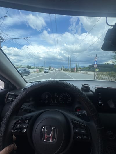 The image is taken from the interior of a car, looking forward through the windshield. The car appears to be in motion, driving on a road with multiple lanes and overhead power lines. The sky is cloudy with patches of blue.
In the foreground, the steering wheel and dashboard are visible. The steering wheel has a textured grip and controls on the left and right sides. The dashboard includes a digital display showing the speed as 59, likely kilometers per hour, and other vehicle information. The central infotainment screen is also visible.
On the road, there are other vehicles, including a dark-colored SUV in the left lane and a white van further ahead. There are road signs visible, including one indicating a height limit of 3.0 meters and another with a speed limit of 20. The environment appears to be suburban or rural, with some buildings and vegetation visible in the distance.
The overall scene suggests a typical driving experience on a cloudy day, with the car navigating a road with traffic and infrastructure. The specific location in Dobrovskoye Rural Settlement, Ukraine, is not visually confirmed by any landmarks or signage within the image.