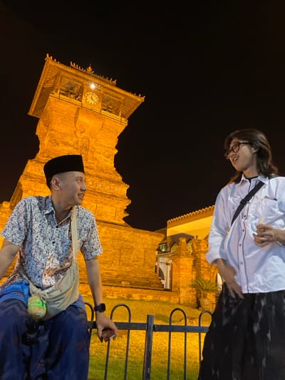 At night in Kauman, Indonesia, two individuals are positioned in the foreground against the brightly illuminated Kudus Menara Mosque clock tower and an adjacent mosque building. On the left, an adult male wears a black peci, a patterned short-sleeved shirt, and a blue sarong, seated on a black metal fence. He looks towards the right, subtly smiling, with a dark smartwatch on his left wrist and an object in his right hand. On the right, an adult individual with shoulder-length dark hair and glasses wears a white long-sleeved shirt with dark piping and dark patterned wide-leg pants, standing and facing left. They are smiling with an open mouth, holding a small white object in their left hand. Both individuals appear to be interacting. The Kudus Menara Mosque clock tower, constructed of reddish-brown brick with a multi-tiered roof and a visible clock face, dominates the background, strongly lit by warm, artificial light against the dark night sky. A strip of illuminated green grass lies behind the fence.