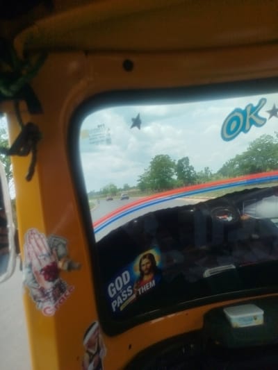 A view from inside a yellow auto-rickshaw in Ukwulu, Nigeria. The vehicle has stickers on its interior, including religious imagery, like Jesus and the phrase "God Pass Them". Through the front windshield, a road is visible with cars and a motorcycle. The sky is overcast.