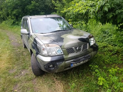 A camouflage-painted Nissan X-Trail SUV is parked on a grassy, dirt track in a wooded area. The vehicle is positioned at an angle, facing slightly towards the viewer's left. Green foliage from trees and bushes surrounds the car, with some branches extending over the hood. The car's headlights are on. The license plate is visible, bearing the inscription "LCA 029" and the letters "LT" in a blue rectangle, indicating it is a Lithuanian license plate. The ground around the vehicle is covered in grass and dirt. The lighting suggests it is daytime.