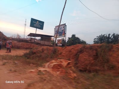 The image displays an outdoor scene in Umudioka, Nigeria, under what appears to be late afternoon or early evening light. The foreground features a reddish-brown dirt path or unpaved road with an adjacent embankment of similar earth, sparsely covered with vegetation. Two individuals, slightly out of focus, stand on the dirt path on the far left; one is discernible wearing clothing with red and orange patterns.
In the midground, a dark green rectangular billboard on a pole is visible, bearing white text that reads "MARANATHA COLLEGE," "UMUDIOKA," and a phone number "0803 891 8230." Behind this billboard, a simple, open-sided structure with a dark, possibly corrugated roof is present. To the right, a second, more colorful billboard is mounted on another pole, displaying indistinct advertisements. A tall, slender wooden pole leans diagonally across the scene from the lower center towards the upper right. Multiple electrical or communication lines traverse the sky overhead. A metal lattice tower is faintly visible in the distant background.
The background consists of dense green foliage, including numerous palm trees on the far right. The sky is light blue with scattered white clouds, showing a subtle gradient of warmer tones near the horizon. The overall image has a slight blur, particularly affecting the subjects and distant objects. A "Galaxy A52s 5G" watermark is present in the lower-left corner.