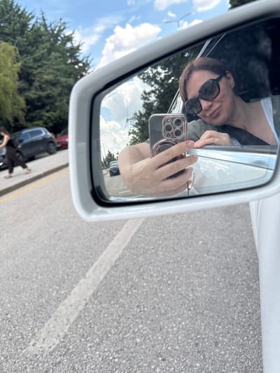 This media file captures a selfie taken by a person inside a car, reflected in the side mirror. The person, wearing sunglasses and a green top over a white shirt, is holding a smartphone with a triple-lens camera. The background visible in the mirror shows a cloudy blue sky and green trees, suggesting an outdoor setting. Reflected in the mirror is also the road with a white line, indicating the car is on a road. The overall mood seems casual, possibly during a drive. The image is oriented vertically. There is no visible text in the image.