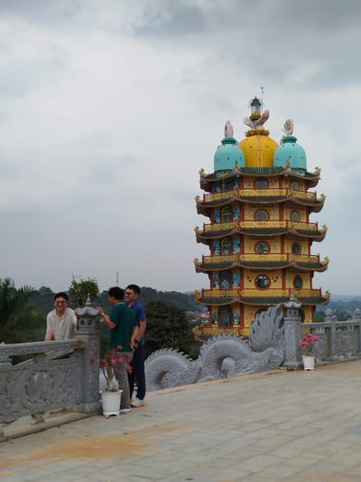 Three men stand on a stone platform near a pagoda-style structure. The pagoda is tall, multi-tiered, and brightly colored with yellow, gold, and blue accents. A decorative stone dragon runs along the edge of the platform. The men are casually dressed and appear to be conversing. The background shows a cloudy sky and a somewhat distant view of trees and possibly other buildings. The location is Thiện Chí, Vietnam.