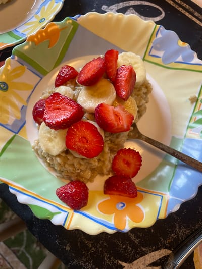 An overhead shot shows a bowl of oatmeal topped with sliced strawberries and bananas. The bowl sits on a decorative plate with a floral pattern. A fork rests on the plate to the right of the bowl. The scene appears to be indoors, likely in a home setting, as indicated by the presence of a table and tableware. There is no identifiable context to place this image in the city of Турово, Russia.