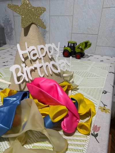 A birthday party setup is displayed on a table covered with a patterned cloth. In the foreground, a pile of deflated balloons in blue, pink, and yellow lies haphazardly. Behind the balloons, a beige cone-shaped party hat is decorated with white "Happy Birthday" lettering and a glittery gold star at the top. Further in the background, a toy tractor, primarily green with red wheels, is positioned on the table. The environment appears to be indoors, possibly a domestic setting. There is no indication of the city of Nerudstal, Ukraine, or any other specific location information within the visual elements.