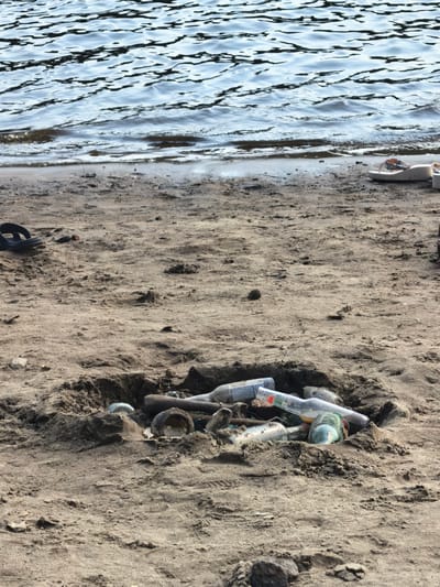 The image depicts a sandy shore of a body of water, likely a river or lake, as indicated by the rippling surface in the background. The foreground is dominated by sand with some scattered debris. Notably, there is a pit in the sand filled with several glass bottles, some of which are partially buried. Additionally, a pair of black flip-flops can be seen on the sand to the left, and a pair of white flip-flops are visible on the right. The overall scene suggests a location in the city of Sysert, Russia, perhaps a recreational area that has been littered. There are no people visible in the image.
