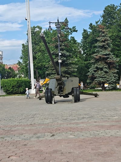 The image depicts an anti-tank gun in the center of a brick-paved public square. It is an overcast day with blue sky and scattered clouds visible. The gun is angled upward towards the sky. Behind the gun, there are trees, streetlights, and white flagpoles. A woman and a small child are walking near the gun. In the background, other people are seen walking in the square, and there is a glimpse of nearby buildings. The location is likely Noginsk, Russia, as suggested by the prompt.