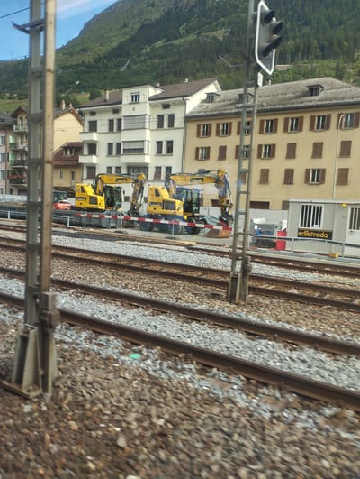 The image captures a scene in Airolo, Switzerland, focused on a construction site adjacent to train tracks. In the foreground, multiple sets of railway tracks run horizontally across the frame, with a utility pole positioned on the left. Behind the tracks, two yellow excavators are visible, one with its boom extended. A red and white construction barrier is in place in front of them. Further back, a light-colored modular building with the signage "edilstrada" is present. Several residential buildings, characterized by their light facades and brown shutters, form the background. A steep, tree-covered mountain rises behind the buildings, with a clear blue sky above. The overall impression is one of ongoing construction work within a semi-urban mountainous environment.