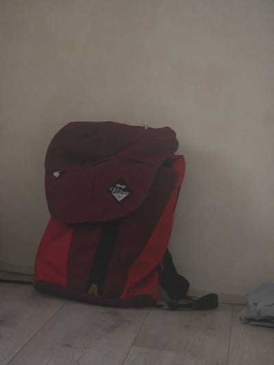 This media file shows a maroon and red backpack leaning against a light-colored wall, with a wooden floor visible to the right. The backpack has a distinctive logo on it, a diamond shape with the word "Nitro" written in script. The backpack appears to be made of a durable material, possibly canvas or nylon, and has a drawstring closure. A black strap is visible extending from the backpack and hanging down towards the floor. To the upper right of the backpack, a piece of light-colored fabric, possibly clothing, is hanging. The lighting in the image suggests it was taken indoors, potentially in a room or hallway. The overall impression is of a casually placed item in a domestic setting. The provided location context is Avtury, Russia, but there are no specific visual cues in the image to confirm this location. There is no discernible activity or event taking place, and the image conveys a sense of stillness.