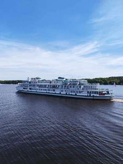 A multi-deck river cruise ship, identified by the Cyrillic text "АФАНАСИЙ НИКИТИН" (Afanasiy Nikitin) painted in blue on its white hull, navigates a wide body of water in Nosovo, Russia. The vessel features a green stripe running along its length and green accents on its upper structures, which include multiple windows and open deck areas. A Russian tricolor flag (white, blue, red) flies from the ship's stern. The ship is in motion, creating a visible wake behind it and generating ripples across the dark blue water surface. Both the left and right banks of the waterway are lined with dense green forests. The sky above is bright blue with scattered, wispy white clouds. No people are visible on the ship or in the surrounding landscape.