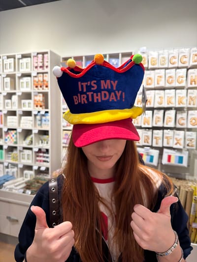 A woman is wearing a red and blue birthday hat with orange text that says "It's My Birthday!" She is giving two thumbs up. She has long red hair and is wearing a black jacket over a white shirt with a red collar. The background shows retail shelves stocked with various items, including letters and cards. The location is a store in Spreitenbach, Switzerland.