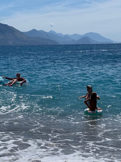 In the city of Lastva Grbaljska, Montenegro, two individuals are in the water. One person, wearing a red long-sleeved shirt and pink swim shoes, is sitting in an inflatable tube. Another person, a child, is also in an inflatable tube, facing away from the camera. A third person, wearing a dark swimsuit and a grey cap, is standing in the water next to the child. The water is a vibrant blue with whitecaps, and mountains are visible in the background under a clear sky. The shore is visible in the foreground with foamy waves.