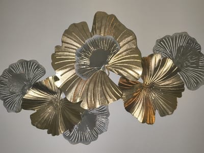 A metallic wall decoration is affixed to a plain, light-colored wall in the city of Liège, Belgium. The decoration consists of a cluster of approximately ten to twelve stylized, fan-shaped leaf elements. These elements vary in size and are primarily golden and silver-grey in color. The individual leaves display distinct radial vein patterns and irregular, scalloped edges, resembling Ginkgo biloba foliage. The golden elements exhibit a reflective metallic sheen, ranging from a bright yellow-gold to a darker, coppery-gold hue. The silver-grey elements range from highly reflective silver to a duller, opaque grey with visible striations. The elements are arranged in an overlapping, three-dimensional configuration, creating shadows on the wall behind and beneath the sculpture, indicating depth. No individuals or actions are depicted in the scene.