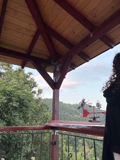 An outdoor scene is captured from a low angle, looking up at the wooden roof of a gazebo. The roof is constructed with light-colored wooden planks and supported by dark red wooden beams. Several metal brackets are visible, securing the beams. In the mid-ground, a black metal railing with decorative scrollwork lines the edge of what appears to be a balcony or deck. Beyond the railing, a lush green forest covers a hillside that slopes down and away from the viewer. A weather vane with a witch figure on a broomstick and windmill blades is prominently displayed on the railing. A portion of a person's dark curly hair is visible on the right side of the frame, indicating their presence. The sky above is a pale blue with wisps of pinkish clouds, suggesting either dawn or dusk. The location is identified as Pechinska, Bulgaria, though this context is not visually apparent from the image itself.