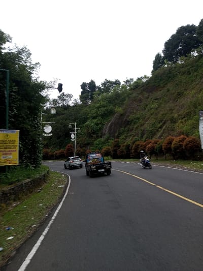 This image depicts a multi-vehicle traffic scene on a paved, two-lane road under an overcast sky, located in Phoxay, Laos.

The dark asphalt road curves gently to the right in the foreground and midground. It is delineated by a solid white line on the left edge, separating it from a narrow grassy shoulder and a concrete barrier, and a double solid yellow line marking the center for two-way traffic.

Three vehicles are visible, traveling away from the viewer in the primary lane: a silver compact SUV leads, followed by a dark blue pickup truck with its bed loaded with large, spiky, yellowish fruits. To the right of these vehicles, a single individual on a dark-colored motorcycle, wearing a helmet, also travels away from the viewer.

On the left side of the road, dense green vegetation forms a wall of trees and bushes. Partially visible within this foliage is a yellow rectangular sign displaying dark text. Three ornate white lampposts with circular white emblems are spaced along the left side. An indistinct dark object, possibly a traffic light, hangs from an overhead cable amidst the trees.

The right side of the road is bordered by a uniform row of reddish-brown ornamental shrubs. Beyond these, a steep hillside rises sharply, covered with green scrub, trees, and exposed rock formations. The sky above is uniformly light grey and overcast.