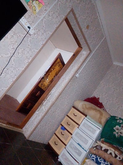 The image captures an indoor scene, possibly a room in a residence located in Кугейский, Russia. The room features patterned wallpaper with a floral or damask design. In the foreground, there is a set of stackable plastic drawers, some of which are tan and have a woven texture, while others are white and have a perforated design. Adjacent to the drawers, there are rolled-up blankets and fabrics, including one with a green floral pattern and another with a blue and white patterned design.
Further into the scene, a wooden doorway with a decorative inlay is visible. Inside the doorway, there appear to be shelves or drawers within a wooden cabinet. A power strip with an attached electrical cord is mounted on the wall near the ceiling. The flooring consists of dark, tiled squares. The overall lighting suggests an interior, possibly dimly lit, environment. There are no people or animals visible in the image.