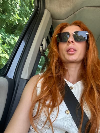 A young woman with long, wavy red hair and sunglasses is inside a vehicle, sticking her tongue out. She is wearing a white top with gold buttons and a black seatbelt. The background shows blurred green trees and foliage, suggesting a rural or natural setting. The image does not provide any specific information to locate it within Medinski Krš, Montenegro.
