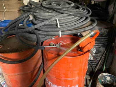 This image, taken in Babawa, Nigeria, depicts an indoor storage or workshop environment. In the foreground and midground are two prominent orange metal drums. The drum on the right features a visible label indicating "Energizer EP 320" and has an orange, hand-operated pump with a brass-toned pipe inserted into its top opening. A large quantity of black, corrugated flexible conduit, some coiled and some loosely arranged, rests atop both orange drums and extends upwards into the background. Several coils of this conduit are secured with white ties. A section of this black conduit runs from the left side, over the left orange drum, and connects to a metallic fitting on the right orange drum. Partially visible behind the conduits on the left is a dark blue plastic barrel. Another dark metal drum is partially discernible behind the right orange drum. In the upper left background, stacked cardboard boxes tied with white string are visible, alongside other miscellaneous items, suggesting a cluttered storage area. No individuals are present.