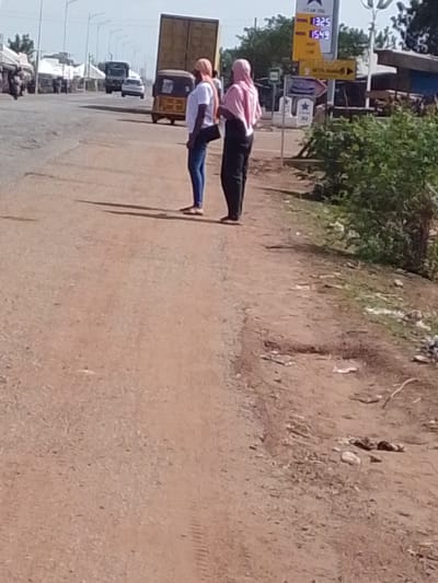 The image shows two people standing on a dirt shoulder of a road in Dungu, Ghana. The person on the left is wearing a white t-shirt, blue jeans, and sandals, with a black purse slung over their shoulder. The person on the right is wearing a pink hijab and black pants.

Behind them on the road, there is a yellow motorized tricycle, a white car, and a truck. In the background, there are more vehicles and buildings. To the right of the people, there is a gas station sign displaying the price of gas at 1325 and 1549. There is also a green bush.

The road surface is a mix of dirt and gravel. The scene appears to be during the day in sunny weather, judging from the shadows cast by the people.