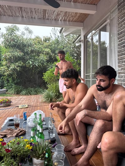 The image captures three men relaxing on an outdoor wooden deck or patio, surrounded by lush greenery. Two men are seated closely together on a lounge or cushions in the foreground, both shirtless with beards, appearing relaxed and engaged in conversation or contemplation. The man closest to the viewer has hairy arms and legs and wears a silver bracelet. A third man stands or crouches further back to the left, also shirtless, holding a smartphone, and wearing light-colored shorts.
The immediate foreground features a black outdoor table laden with remnants of a social gathering. Several empty green glass bottles, some identifiable as "Corona" beer, are clustered in a glass pitcher with ice, while others stand upright. Numerous clear plastic cups are scattered across the table, some empty, others containing liquid. A glass with a dark beverage and a blue and white can marked "XX" are also visible. One man holds a lit cigarette. A white plastic bag, a brown cloth or jacket, and a white electronic device with a blue cable are also on or next to the table. Potted plants with vibrant flowers and a succulent provide decoration.
The setting features a wooden deck floor and is enveloped by dense green trees and bushes, indicative of a warm climate like Hod HaSharon, Israel. Above, a modern structure with a glass roof or skylight is supported by white beams, attached to a brick wall. A security camera is mounted on one of these beams. Bamboo blinds are visible on the left. The lighting is soft and diffused, suggesting an overcast day or late afternoon. The overall mood is casual and social.