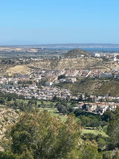 The image depicts an elevated view of a portion of the city of Rojales, Spain, situated near the coast. The landscape includes a mix of residential areas, characterized by densely packed white buildings, interspersed with vegetation. A prominent, somewhat barren hillside rises through the center of the frame, with houses built on its slopes. To the right, a well-maintained golf course is visible, with green fairways and mature trees. The horizon shows the coastline and the Mediterranean Sea, with a hazy blue sky overhead. The overall environment appears arid and sunny.