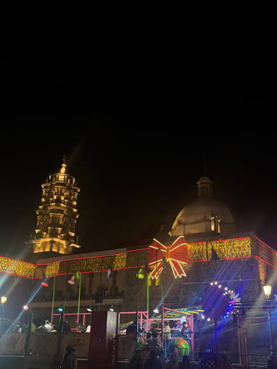 A dark, nighttime scene shows a church dome illuminated on the upper right. Below and to the left is a decorative tower also lit up. The church structure is adorned with Christmas-themed decorations, including golden ribbon-like lights and a large red bow. Part of a fair or carnival ride is visible in the background along with a crowd of people. The photograph seems to be taken during a celebration or event.