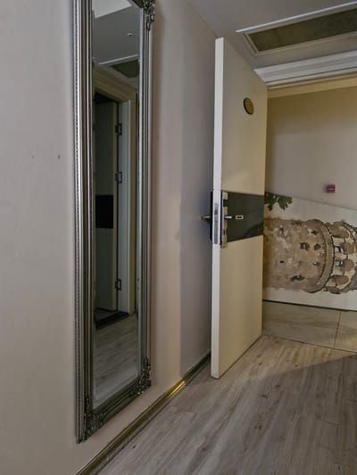 The image depicts an interior scene, likely a hallway, with a large, ornate silver-framed mirror leaning against a pale-colored wall on the left. The mirror reflects a doorway and part of a floor. To the right, a white door stands ajar, revealing a dark-colored panel with a silver handle and lock mechanism. Above the door, a rectangular ventilation grille is visible in the ceiling. The wall to the right of the door features a large, faded mural or wallpaper depicting a circular, stone structure. A small red fire alarm box is mounted high on this wall. The flooring is a light-colored wood laminate. The scene appears to be in a state of partial disrepair or renovation, as suggested by the unfinished mural and the condition of the wall. The location is specified as Bakırköy, Turkey. There are no people or animals present in the image.