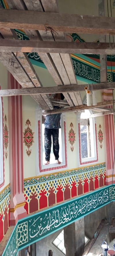 The image displays the interior of a building, likely a mosque, undergoing decorative painting in Kumbang, Indonesia. A single individual is positioned centrally in the mid-ground, standing barefoot on the sill of an open window. This person, wearing a paint-splattered light-colored shirt and dark trousers, is engaged in applying paint or decorative finishes to the adjacent wall.

Above the individual, a temporary scaffold constructed from rough, unpainted wooden planks is visible, with some planks showing paint splatters and protruding nails. Two white paint rollers and two yellow pipe fittings are suspended from one of these planks.

The walls are extensively decorated with intricate patterns and calligraphy. The upper visible wall section is cream-colored, featuring pink-outlined window frames and symmetrical red, green, and gold ornamental scrollwork surrounding the windows. Below the windows, a multi-colored frieze presents repeating red and brown stylized arch patterns, topped by a band of interwoven blue, green, brown, white, and yellow diamond tessellations. Directly beneath this, a prominent horizontal teal band displays large white Arabic calligraphy. Below the teal band, a green band features smaller white Arabic script interspersed with white geometric star patterns.

In the lower right portion of the image, unpainted grey concrete pillars and exposed wooden planks are visible. Two white paint cans and one blue paint can rest on the floor. Natural light enters the space through the windows.