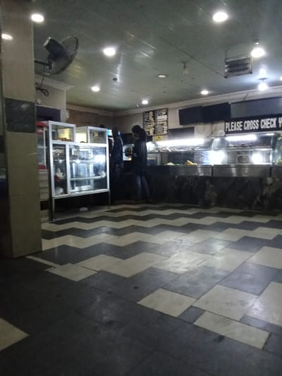 The image shows the interior of a food stall or eatery in Ovwian, Nigeria, located at coordinates 5.503 latitude and 5.799 longitude. There are three people standing near a stainless steel counter, which is filled with food displayed in glass containers. The counter has a sign that reads "PLEASE CROSS CHECK YOUR ORDER". The eatery has a tiled floor with a black and white pattern. The walls are painted in a muted beige color, and the ceiling is made of white-painted panels. There is a ceiling fan in the upper left corner of the picture. There are several fluorescent light fixtures hanging from the ceiling, which are illuminating the scene.  A large, silver, glass-doored cabinet is visible to the left of the counter. There are two other, smaller glass-doored cabinets to the left of the large cabinet. There are some unknown objects stored in the cabinets. The people in the scene are wearing dark clothing and their faces are not visible.