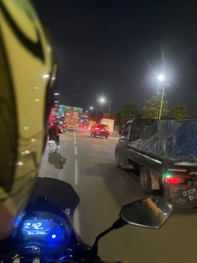 This image was taken at night on a road in Sawotratap, Indonesia. The perspective is from a motorcycle, with the rider's yellow and black helmet and a portion of the handlebars and dashboard visible in the foreground. The dashboard displays a speed of 42 km/h, a battery level of 65%, and other trip data.
The scene depicts traffic on a multi-lane road. Several vehicles are present, including a dark-colored truck with a blue tarp covering its cargo, a red car, and other vehicles partially obscured by darkness and distance. A motorcycle with a rider wearing a red shirt and dark pants is visible in the middle lane. Traffic lights are red in the distance, indicating a possible traffic stop. Streetlights illuminate the road and the surrounding environment, which includes trees and overhead signs. The sky is dark, with a bright light source, possibly the moon or a streetlamp, visible towards the upper right. The road appears to be paved and marked with white lines.