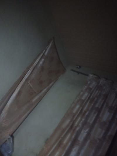 The photograph depicts an interior scene, likely a room, in Tudun Nupawa, Nigeria. The visible components include two sections of draped curtain, with a floral pattern, against walls of a light color. The lighting is dim, obscuring details. A curtain rod is visible near the top of the frame. At the bottom of the frame, a dark mass of what looks like a plastic bag can be found. The perspective is tilted, giving the impression of viewing the corner of a room at an upward angle. The overall setting appears to be a residential space.