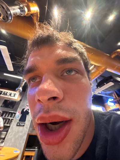 A close-up selfie, rotated counter-clockwise, features a man making a humorous expression by sticking his tongue out widely to the side. He has light-colored eyes, dark, disheveled hair, and stubble on his face. His skin appears somewhat shiny.

The background, largely out of focus, reveals an indoor setting with an industrial design aesthetic, likely a restaurant or bar. Golden-colored industrial piping runs across the upper and left parts of the frame, with a silver metal valve fixture visible in the lower left. Several bright light sources create prominent starburst lens flares. A blue neon light is partially visible in the upper right. In the mid-right, a sign features a stylized caricature of a man in a suit and prominent Chinese text. Parts of a wooden, surfboard-like object are visible on the far right.

The visible Chinese text on the sign reads "本肠先生" (Běn cháng xiānshēng), which translates to "Mr. Sausage" or "Mr. Intestine." Smaller Chinese characters are also present below this text.

The scene appears to be set at night or in the evening, indicated by the interior lighting. The man's playful expression suggests a lighthearted moment. The presence of Chinese text and the industrial decor are consistent with a setting in Minhang District, China.