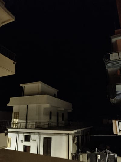 The image shows a nighttime view of residential buildings in Bova Marina, Italy. The foreground features a dark, textured surface that appears to be a wall or railing. To the left, a portion of a white building with a balcony is visible against a dark sky. The central focus is a multi-story white building with a flat roof and balconies. The building has several windows, some with shutters, and at least one visible air conditioning unit. The lower level of this building also has windows and doors. To the right, another building is partially visible, with sections covered by striped awnings. A metal gate with an informational sign is present in the lower right corner. The overall scene is dimly lit by artificial light sources, suggesting it is nighttime.