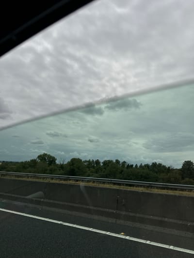 The image captures a view from a vehicle traveling on a highway, likely in or near Ballitore, Ireland, given the context provided. The scene is dominated by a cloudy sky, with a distinct diagonal line cutting across the frame, possibly representing the window of the vehicle or a reflection. Below the sky, a dense line of trees forms a natural barrier. In the foreground, a dark asphalt road with a white lane marking is visible, indicating a roadway. A concrete barrier separates the road from the area with the trees and sky. The overall impression is one of overcast weather and a typical roadside landscape. There are no people or specific actions depicted in the image.