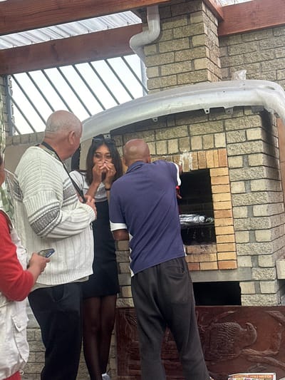 The scene depicts an outdoor gathering in Mitchells Plain, South Africa, featuring four individuals positioned around a brick braai structure. To the far left, a person is partially visible from the chest down, wearing a red long-sleeved top and a light-colored sleeveless garment, holding a black smartphone. Standing to their right, an older male with a balding head and short grey hair wears a white and grey striped knitted sweater over a dark shirt, dark trousers, and a dark lanyard around his neck; he is looking towards the center of the frame. In the center, a young woman with long dark hair, wearing sunglasses pushed up on her head, a light-colored top, a black skirt, and dark sheer hosiery, clasps her hands near her mouth with a visible smile or reaction of amusement. To her right, an older male with a balding head and short dark hair, dressed in a dark blue polo shirt with white sleeve trim and dark trousers, is bent over and facing a light-colored brick braai structure. He is actively tending to items wrapped in silver foil visible on a grate within the braai's lower opening. A light grey car bumper rests horizontally on top of the brick braai structure. Above the scene, a wooden beam supports a translucent corrugated roof, and a white PVC pipe is attached to the beam and extends down the brickwork. In the left background, grey metal railings are visible against a light sky. A dark wooden panel with carved relief art is partially visible below the braai opening on the right, and a partial sign reading "OMBO_ YOU NEED!" is present at the bottom right.