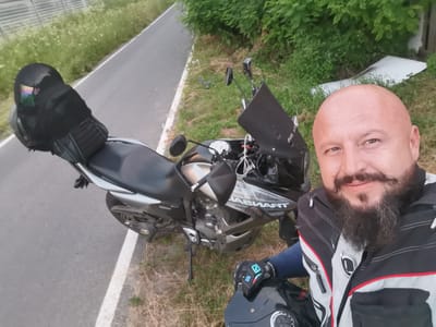 A bald, Caucasian male with a full beard and mustache is taking a selfie next to a black motorcycle on the side of a road. He is wearing a black and grey motorcycle jacket with red trim and black gloves. A black helmet with a reflective visor is on the seat of the motorcycle, and another helmet, also black, is held in his left hand. The motorcycle is parked on the grass next to a paved road with a white line. In the background, there is lush green foliage and a white object that appears to be discarded material. The overall scene suggests a roadside stop during a motorcycle ride. The image was taken in the city of Semypolky, Ukraine.