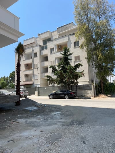 The image depicts an exterior view of a modern, multi-story building with a light-colored facade. The building features numerous balconies and windows, some with shutters. In front of the building, there is a paved area with a gravelly section and some debris scattered on the ground. A black car is parked on the paved area, and behind it, a metal gate is visible. To the left of the building, a tall tree with green foliage stretches upwards, and a palm tree stands prominently in the foreground. The sky is clear and blue, suggesting a daytime scene under fair weather conditions. The scene appears to be in a residential or urban area. No people are visible in the image, and no text is discernible. The orientation of the image is rotated 90 degrees clockwise.